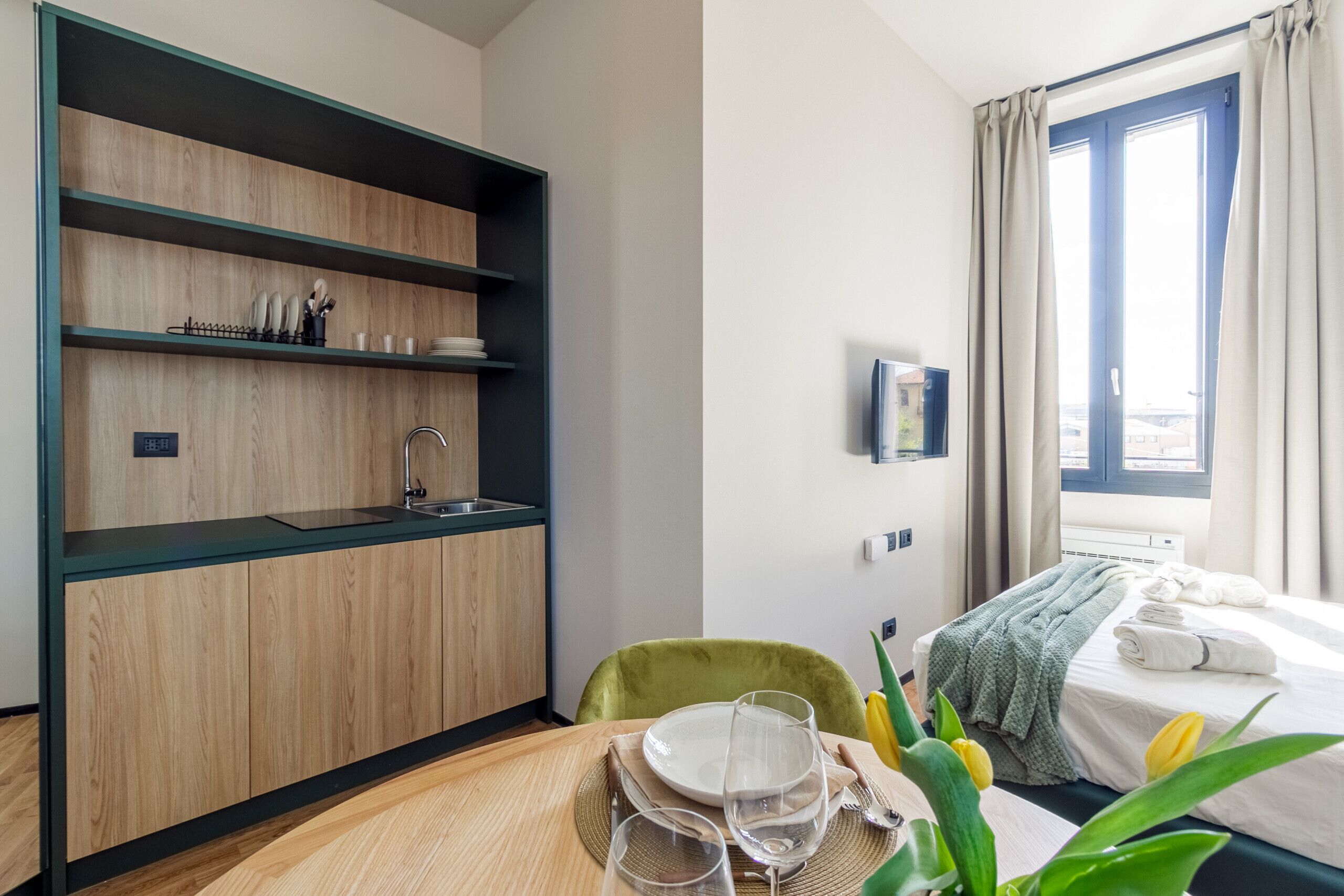 A modern hotel room with a wooden and black shelving unit, a green chair, a bed with a green blanket, and a window with curtains.