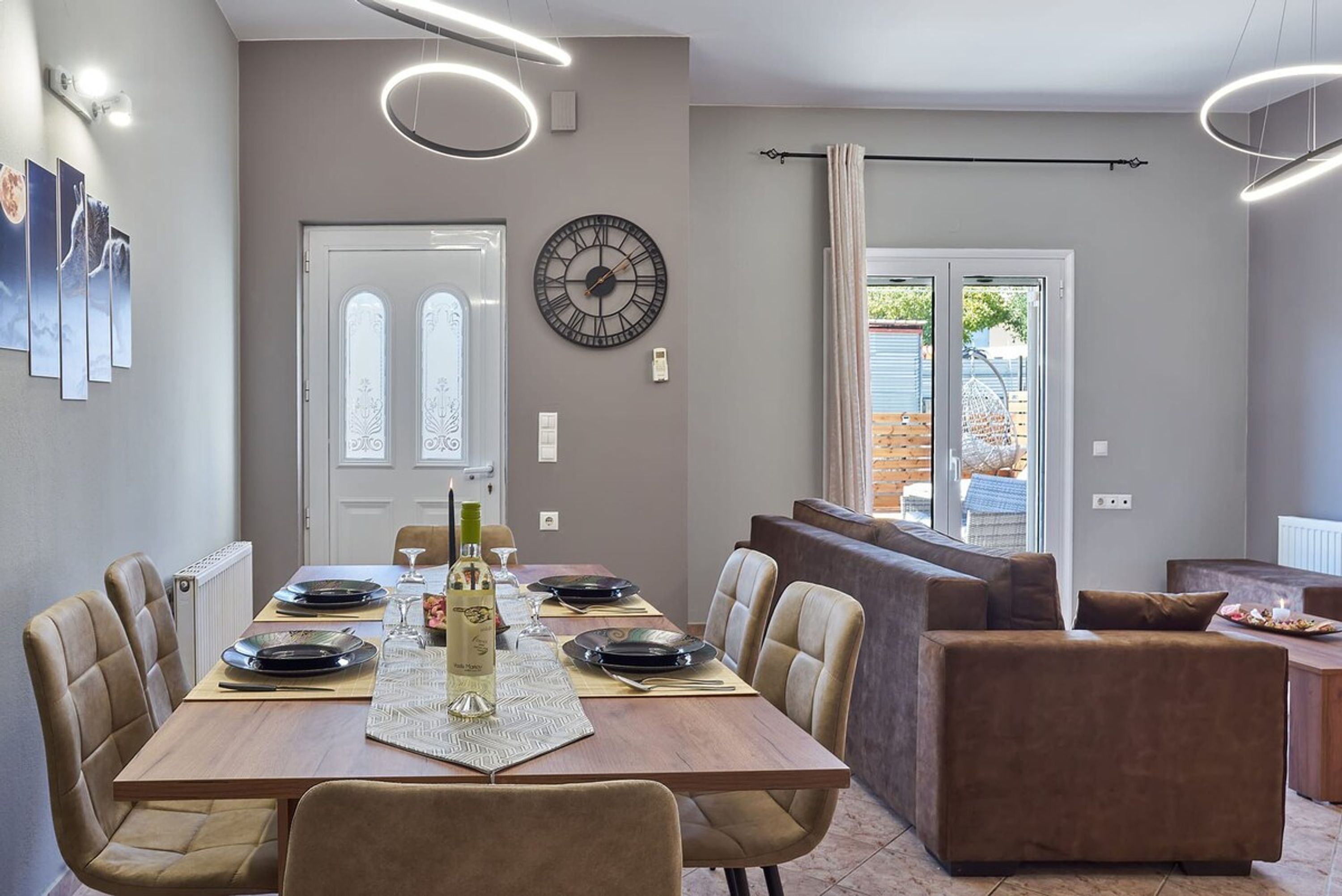 A dining area with a wooden table set for four, beige chairs, a wall clock, and a view of the outside through a glass door.
