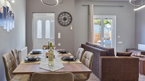 Zona pranzo con tavolo in legno apparecchiata per quattro persone, sedie beige, orologio a parete e vista all'esterno tramite una porta in vetro.