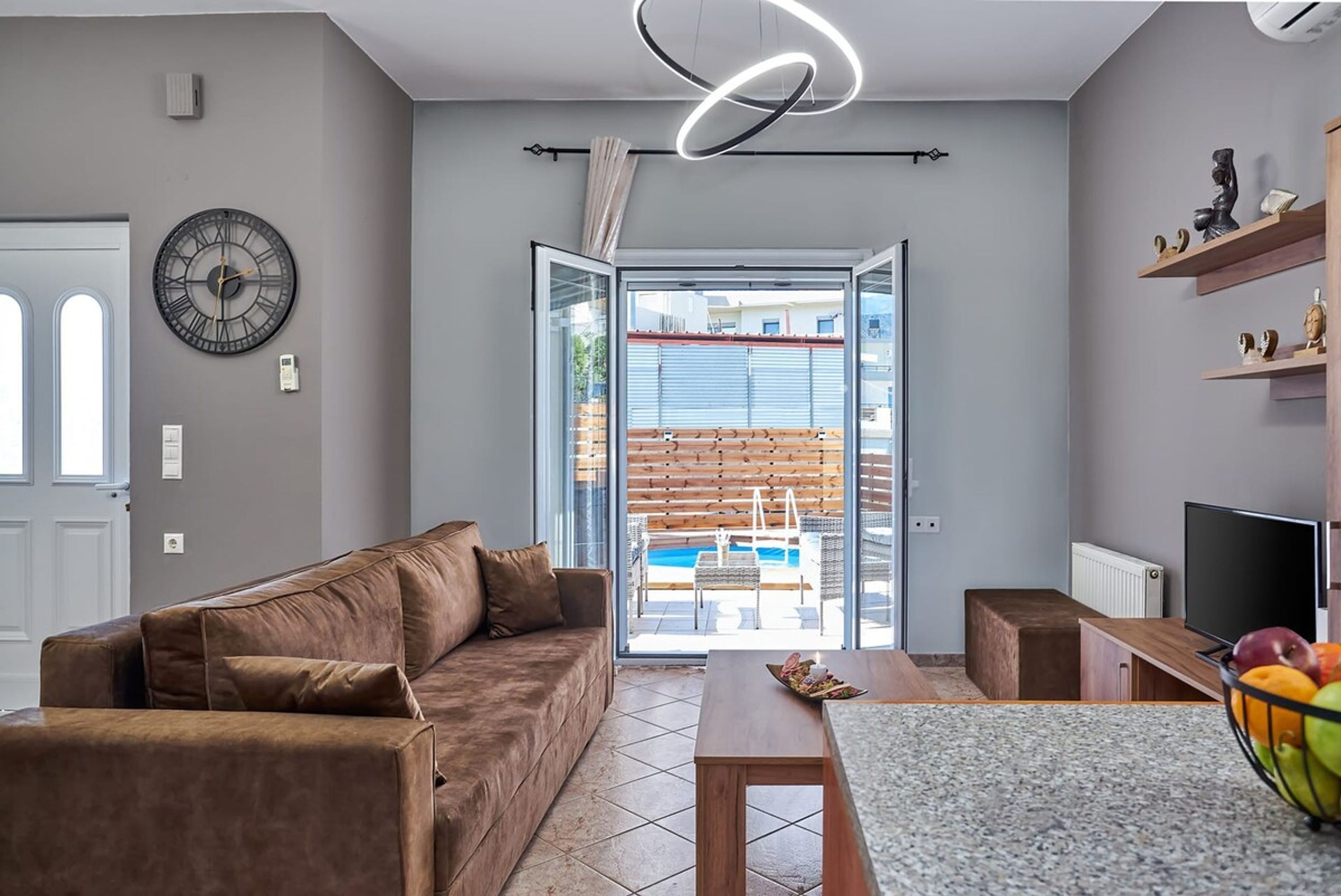 A modern living room with a brown leather sofa, a wooden coffee table, and a TV stand. A large window offers a view of a pool and outdoor seating.