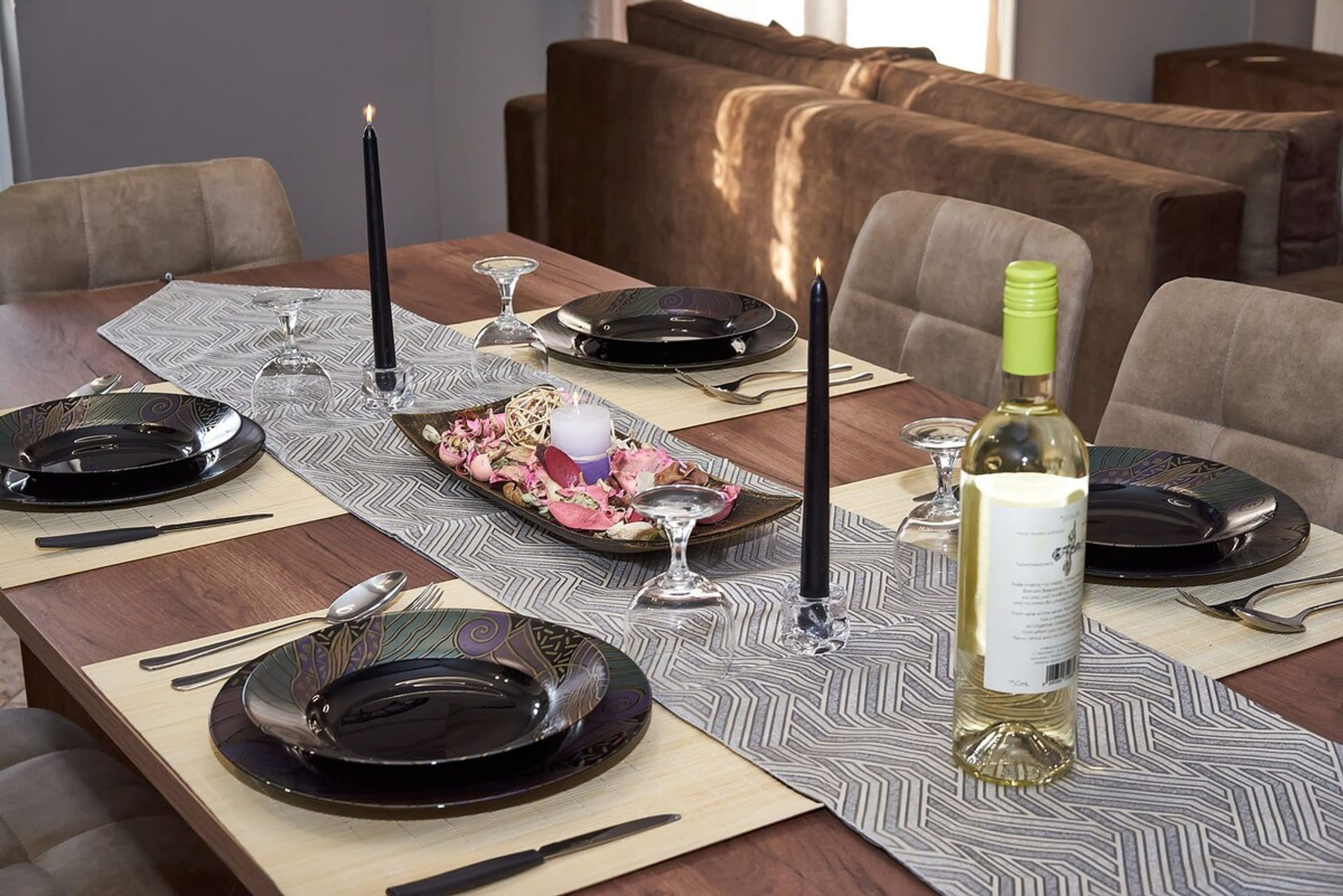 Une table à manger dressée avec des assiettes, des couverts, des bougies et une bouteille de vin, avec un canapé en arrière-plan.