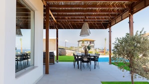 Un patio exterior con una pérgola de madera, una mesa de comedor con sillas y vistas a una casa y piscina.