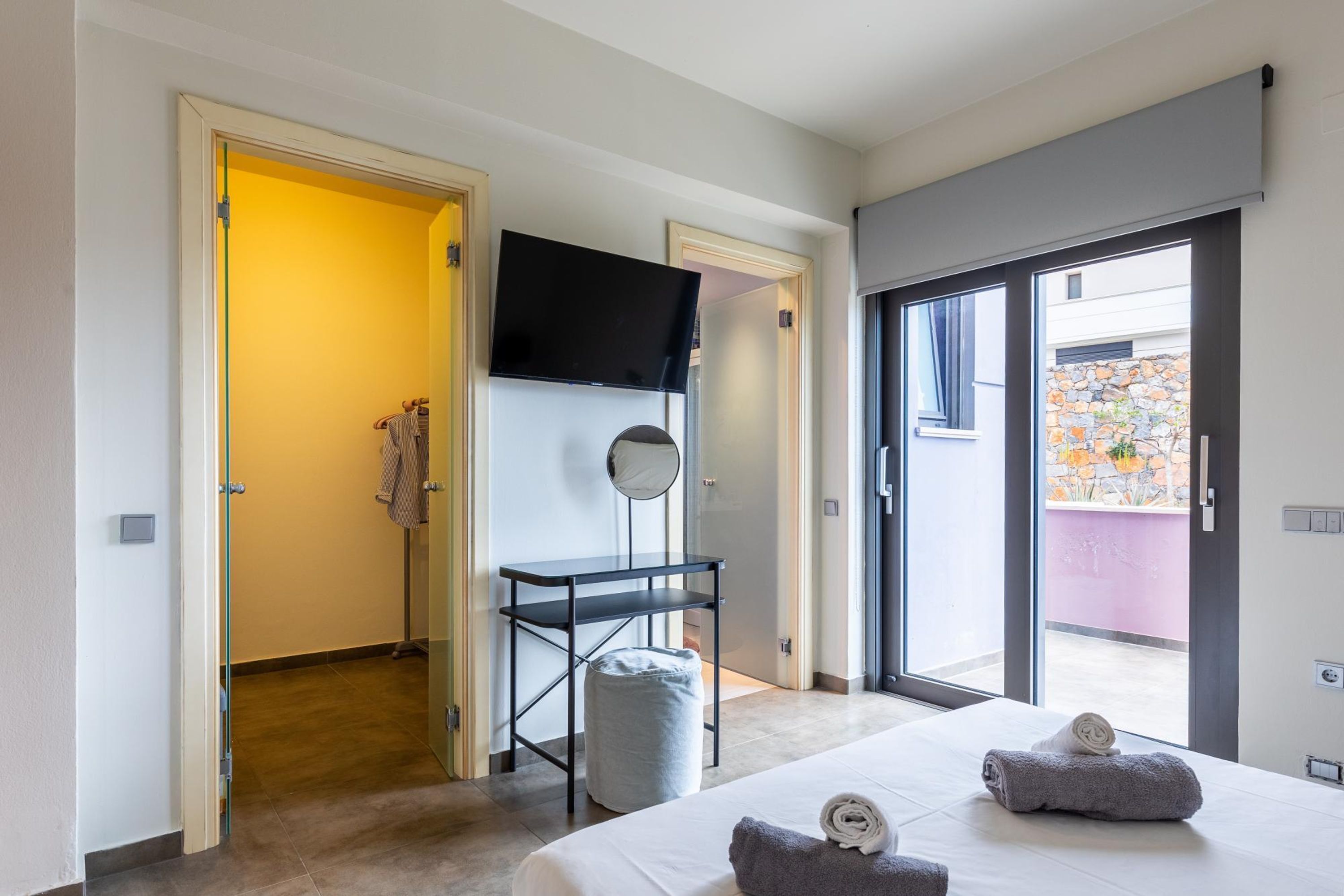 A modern hotel room with a bed, a TV, a small table, and a sliding glass door leading to an outdoor area.