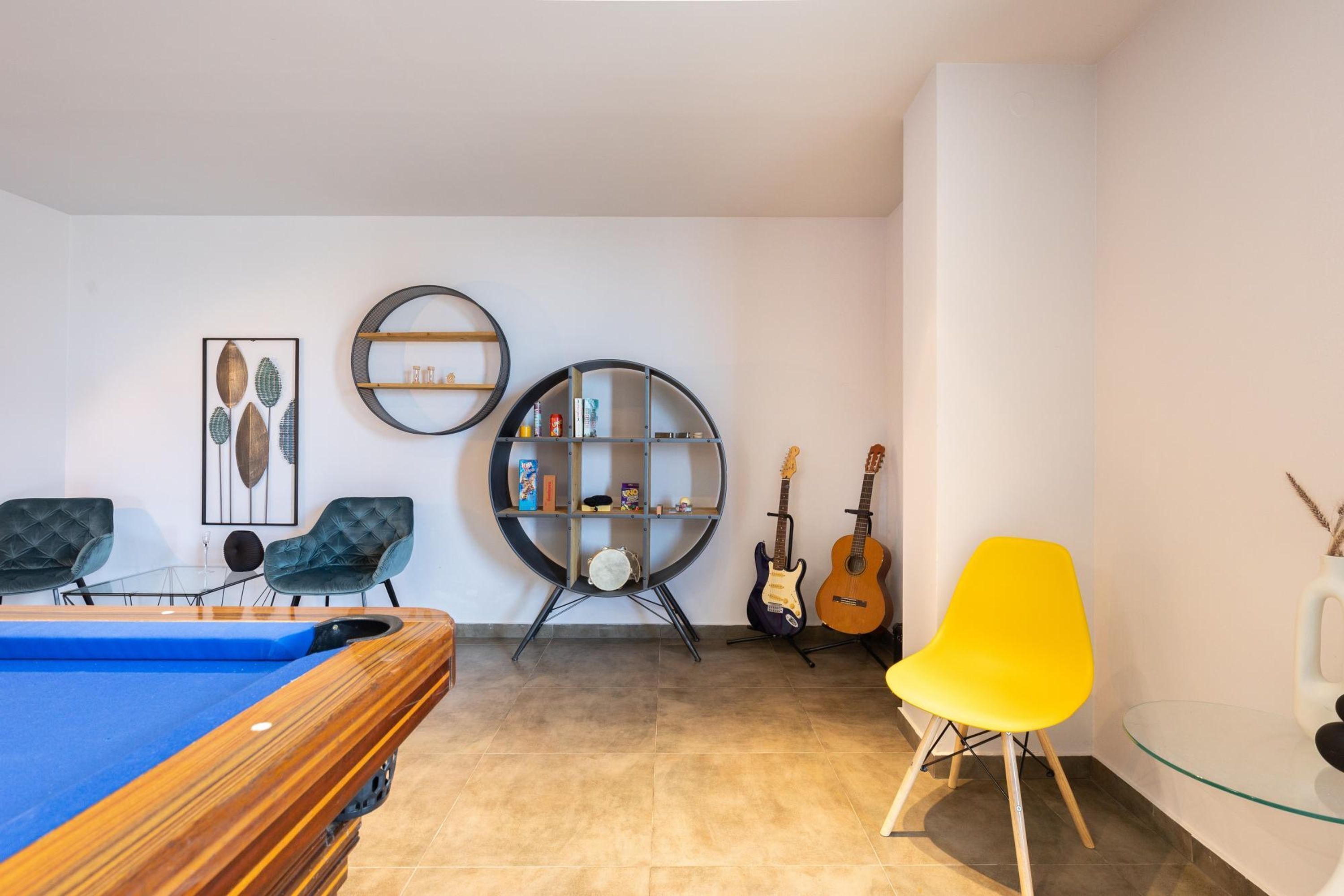 A modern living room with a pool table, two green upholstered chairs, a yellow chair, a glass coffee table, a round bookshelf, and two guitars.