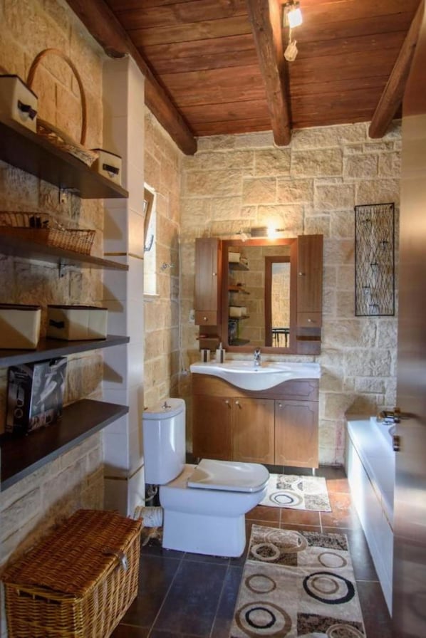 A rustic bathroom with stone walls, wooden beams, a sink, toilet, and shelves with baskets. - Stone and Wood Villa (Hersonissos)