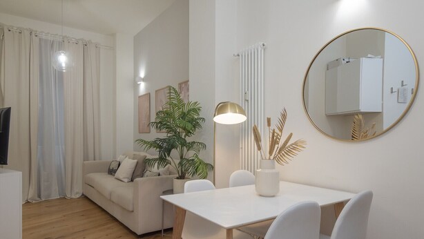 Un salon moderne avec un canapé blanc, une table à manger avec des chaises, une plante en pot, une lampe sur pied, un miroir rond et un radiateur.