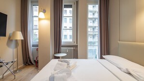 Une chambre d’hôtel avec un lit, une petite table ronde, une lampe et une vue sur les immeubles à travers de grandes fenêtres.