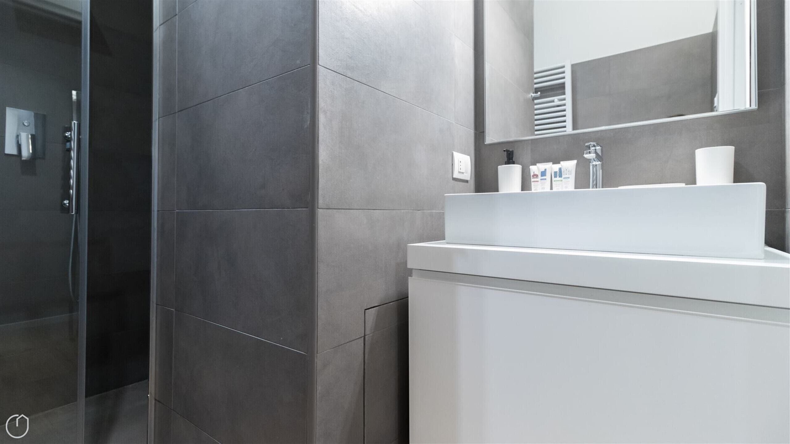 A modern bathroom with grey tiles, a glass shower enclosure, a white sink, and a mirror.
