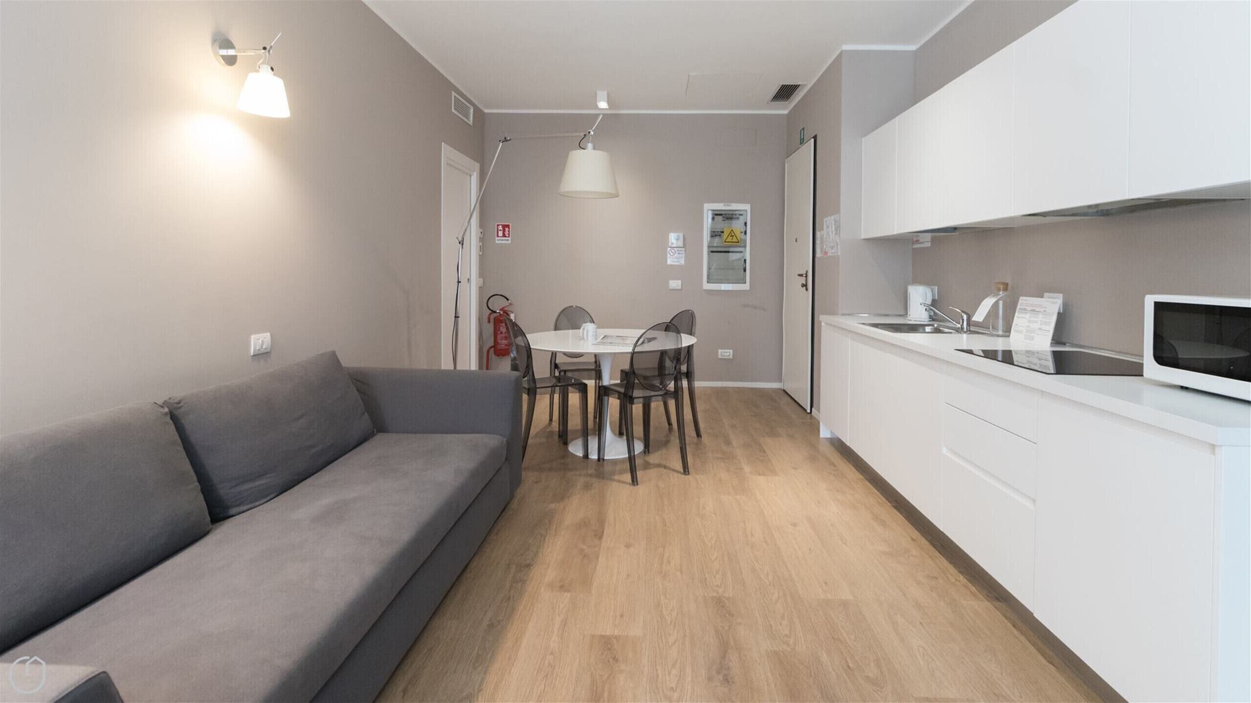 A compact living space with a grey sofa, a round dining table with chairs, a kitchenette with a microwave, and a white cabinet.
