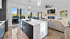 Une cuisine moderne dotée d’un îlot central, un coin repas avec une table et des chaises, un salon avec un canapé et une cheminée, ainsi qu’un ventilateur de plafond.
