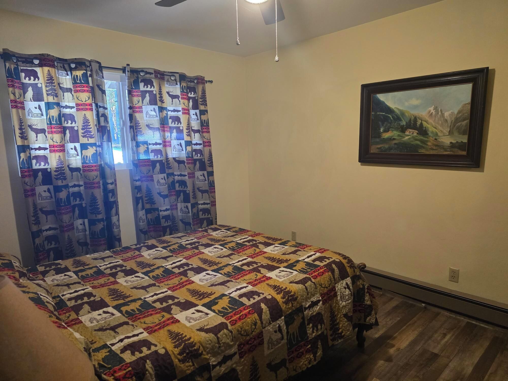 A bedroom with a bed, a painting on the wall, and a window with patterned curtains.