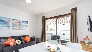 A modern living room with a grey sofa, white dining table, and a view of the outdoors through a glass door. - Home2Book Vibrant Las Américas Apt, Pool Retreat (Playa de la Américas)