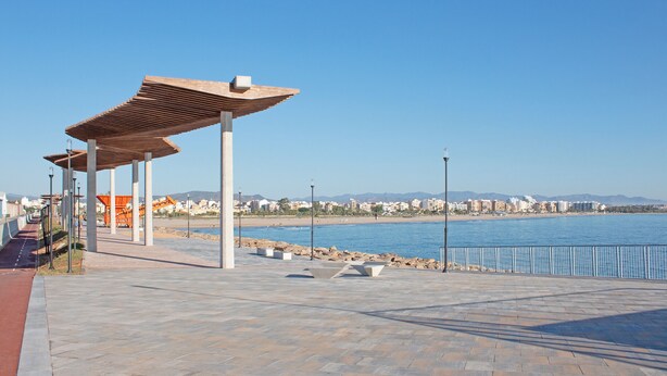 Un paseo marítimo con zona de estar cubierta, un sendero pavimentado y vistas al mar y edificios lejanos.