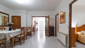 Un comedor con una mesa, sillas y un jarrón de flores. Se observa un armario de madera y una cama con cabecera al fondo.