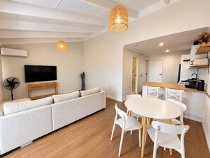 Un soggiorno moderno con divano, televisione, tavolo da pranzo con sedie e angolo cottura con piano di lavoro ed elettrodomestici.
