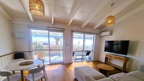 Una sala de estar moderna con un sofá, un televisor y una mesa de comedor con sillas. Se observan grandes ventanales y un balcón.