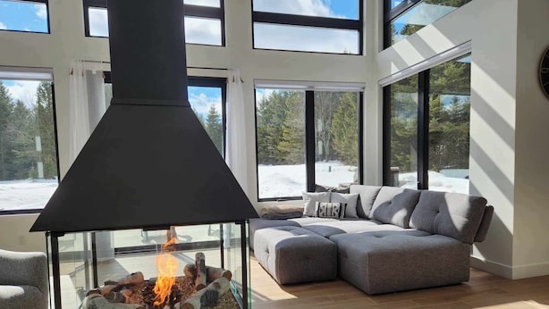 A modern living room with a glass fireplace, a grey sectional sofa, and large windows offering a view of a snowy landscape. - Ô paradis Spa&Massage (Weedon)