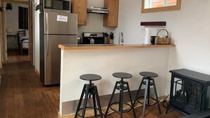 A compact kitchen with a refrigerator, microwave, and stove, wooden cabinets, a small island with stools, and a black wood-burning stove. - Native Spirit Cabin at Base Camp (Dumont)
