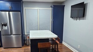 A kitchen with a wooden floor, a built-in island with seating, a stainless steel refrigerator, and a flat-screen TV mounted on the wall.