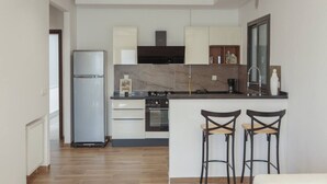 A modern kitchen with a built-in oven, a refrigerator, and a marble backsplash. There are two bar stools and a window with a view of the outdoors.