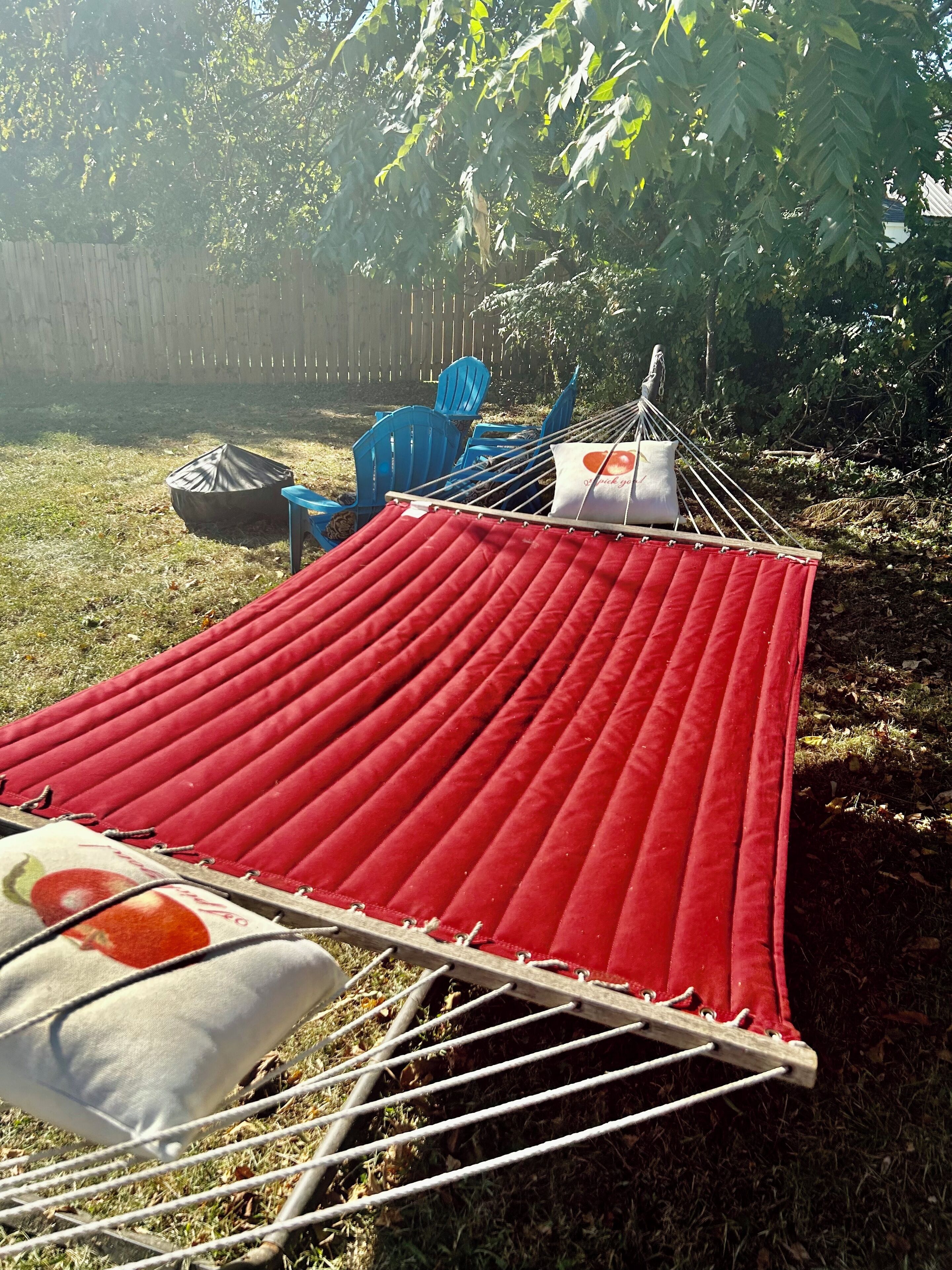 A hammock with a red fabric, a white pillow with a red design, and a blue chair in a backyard.