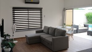 Un salon moderne avec un canapé d’angle gris, un ventilateur sur pied et un coin repas avec des sièges d’extérieur.