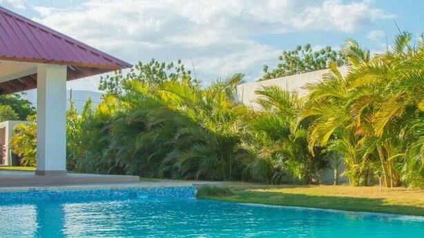 A swimming pool with clear blue water, surrounded by lush greenery and a building with a red roof.
