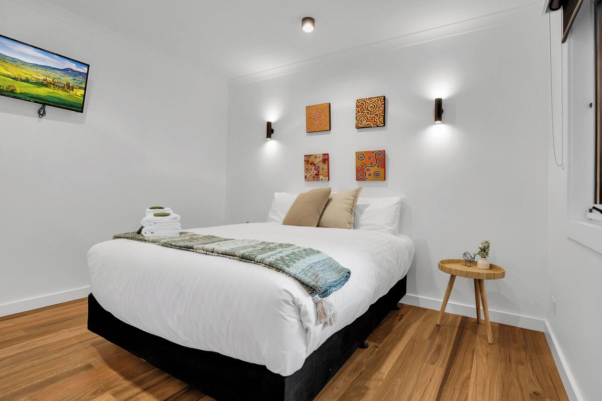 Een net opgemaakt bed met een witte deken en een blauw-witte plaid, een klein houten nachtkastje met een plant, en vier wandlampen.