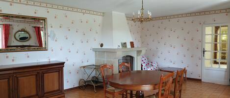 A dining room with a round wooden table, chairs, a chandelier, a fireplace, and patterned wallpaper.