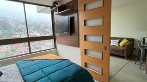 Un dormitorio moderno con una cama grande, un televisor en la pared y una puerta corredera que da a un balcón con vistas a la ciudad.
