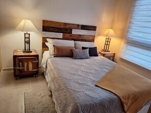 A bedroom with a wooden headboard, a bed with patterned bedding, two bedside lamps, a nightstand, and a window with blinds.