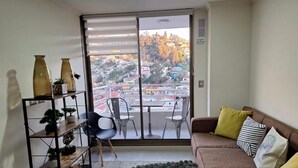 Una sala de estar con un amplio ventanal corredera que da a un balcón, un sofá con cojines, una mesita y una silla.