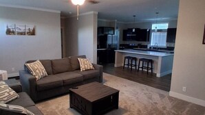 Una sala de estar con sofá, mesa de centro y ventilador de techo. Se ve una cocina con taburetes de bar al fondo.