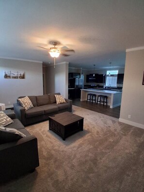 A living room with a sofa, coffee table, and a ceiling fan. A kitchen with bar stools is visible in the background.