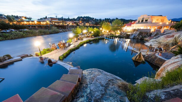 Un spa junto al río con piscina, escalones que bajan al agua y un edificio iluminado al fondo.