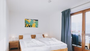 A modern bedroom with a wooden bed, a nightstand with a lamp, a painting on the wall, and a view through a glass door. - Apartments Haus Altenberger - Studio, Lake view & Balcony (Zell am See)