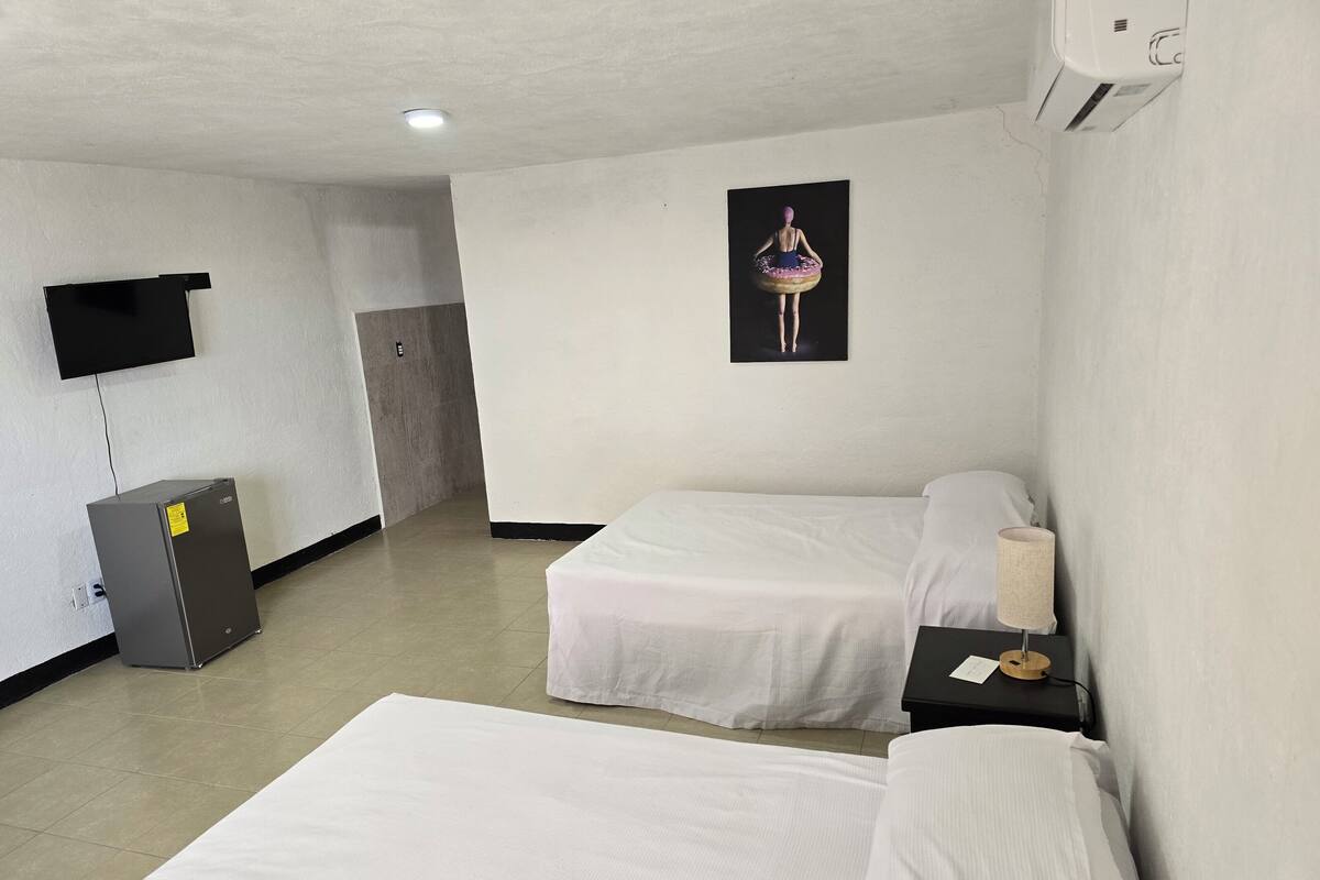 A hotel room with two single beds, a small desk with a lamp, a TV mounted on the wall, and a refrigerator.