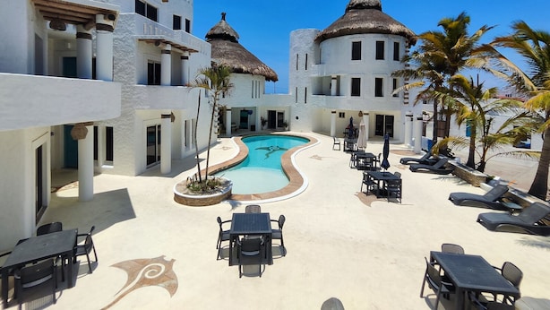 A resort with a swimming pool surrounded by lounge chairs and tables, white buildings with thatched roofs, and palm trees.