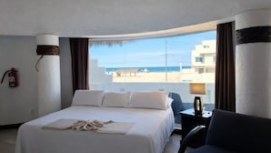 A hotel room with a bed, a chair, a nightstand, a lamp, a fire extinguisher, and a view of the beach and buildings.