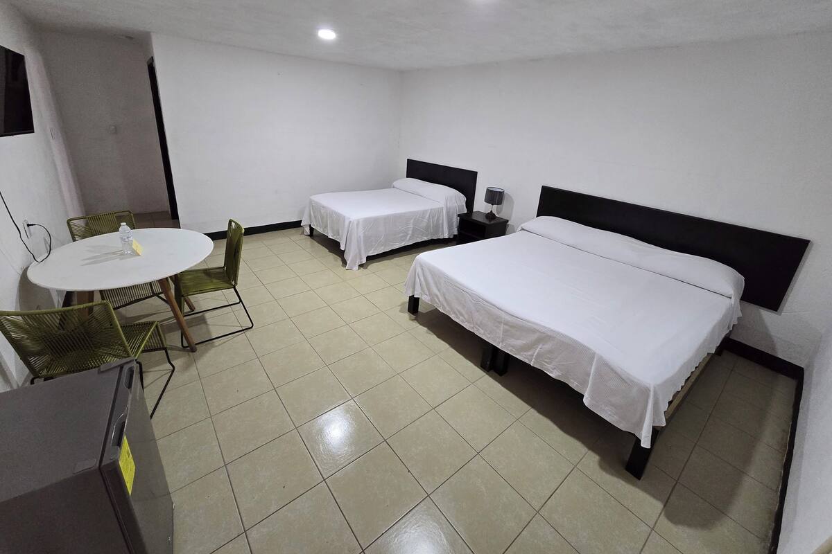 A hotel room with two single beds, a round table with chairs, a small bedside table with a lamp, and a refrigerator.
