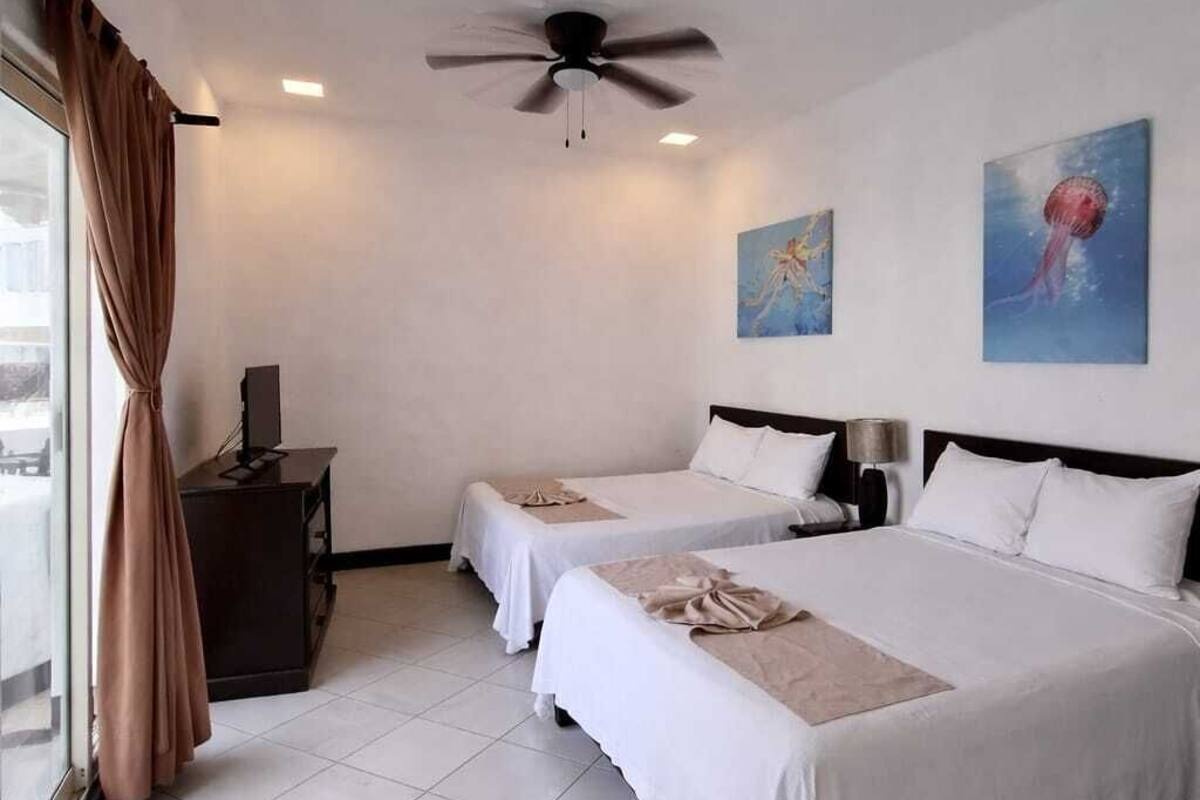 A hotel room with two beds, a ceiling fan, artwork on the walls, and a window with curtains.