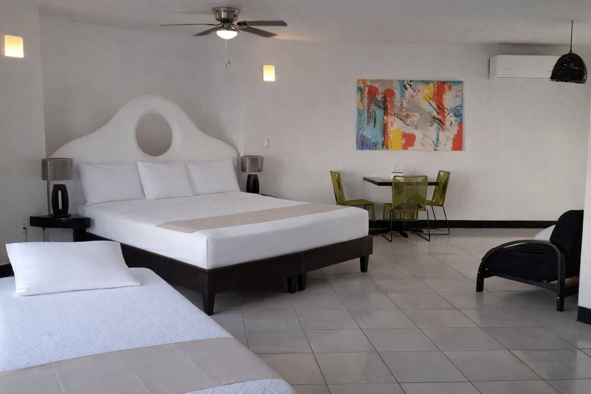 A modern hotel room with a large bed, a desk with chairs, a ceiling fan, and a colorful abstract painting on the wall.