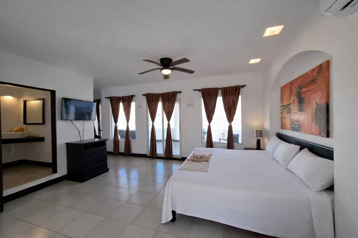 A hotel room with a bed, a TV, a ceiling fan, and a painting on the wall.