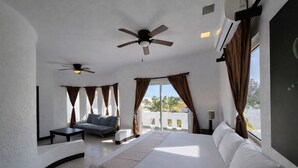 A hotel room with a large bed, a sofa, a small table, two ceiling fans, and a view of palm trees through the window.