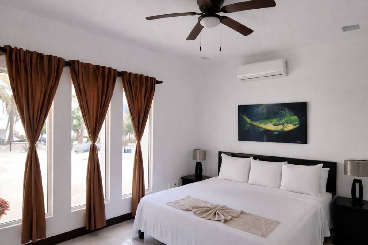A hotel room with a large bed, a ceiling fan, a painting on the wall, and a view of the outside through the windows.