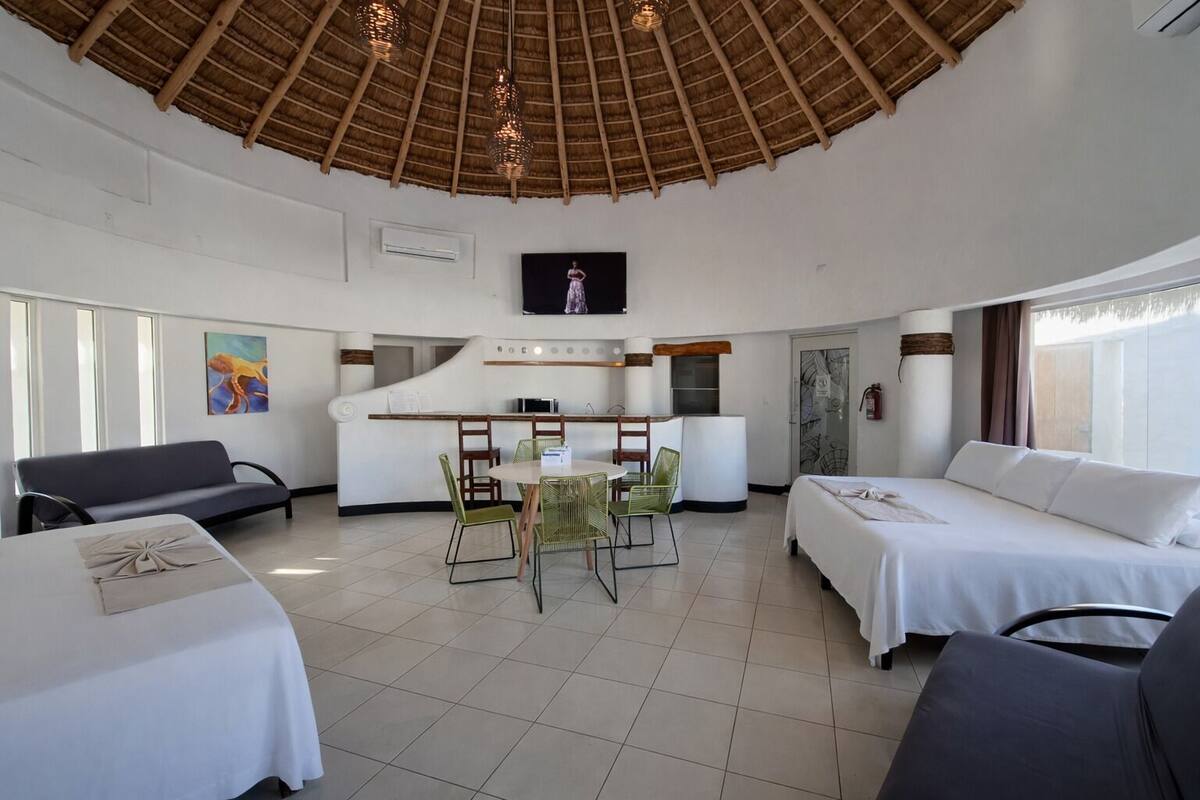 A hotel room with two beds, a dining area with chairs, a TV, and a thatched ceiling.
