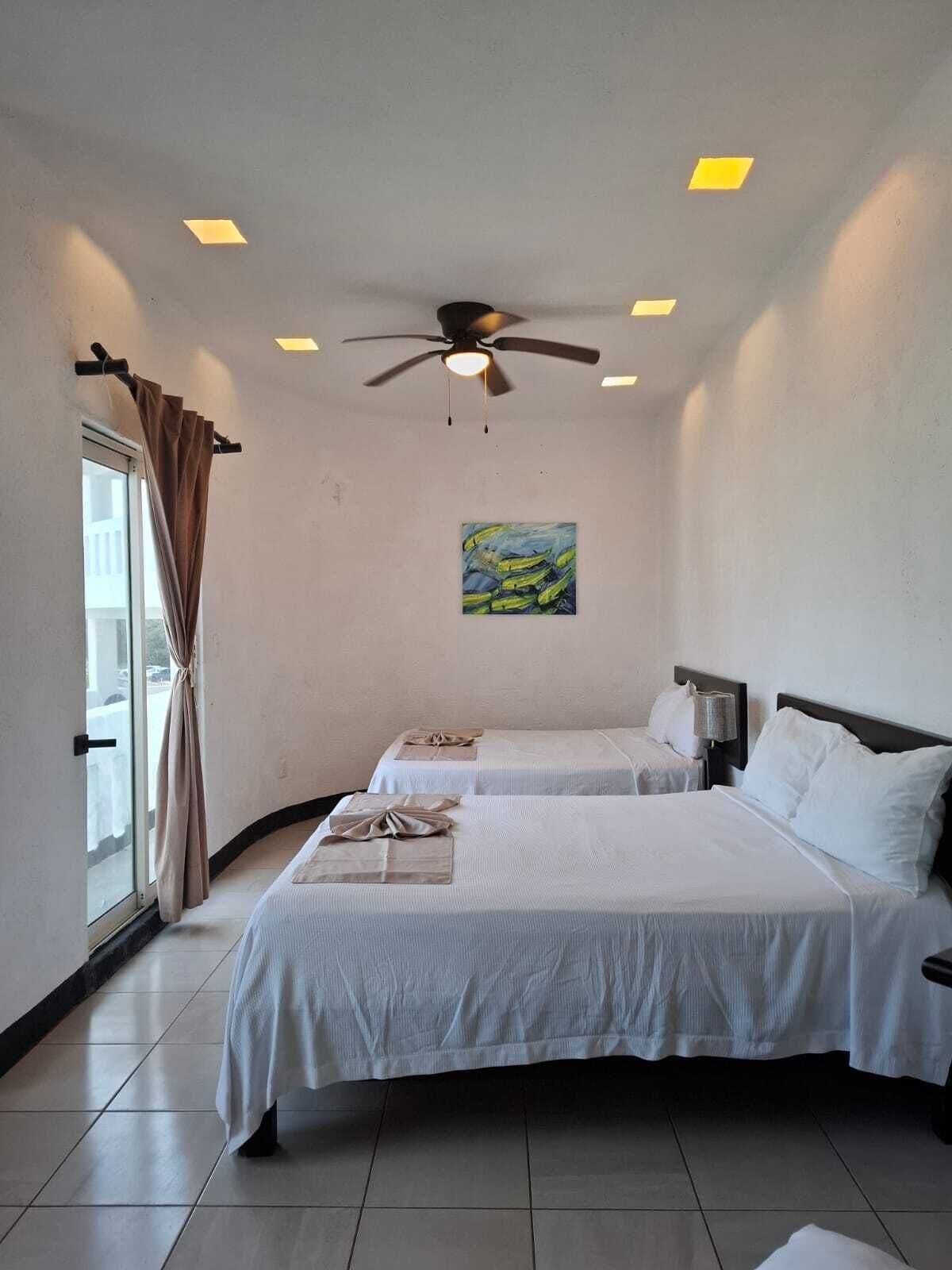 A hotel room with two beds, a ceiling fan, a painting on the wall, and a window with curtains.