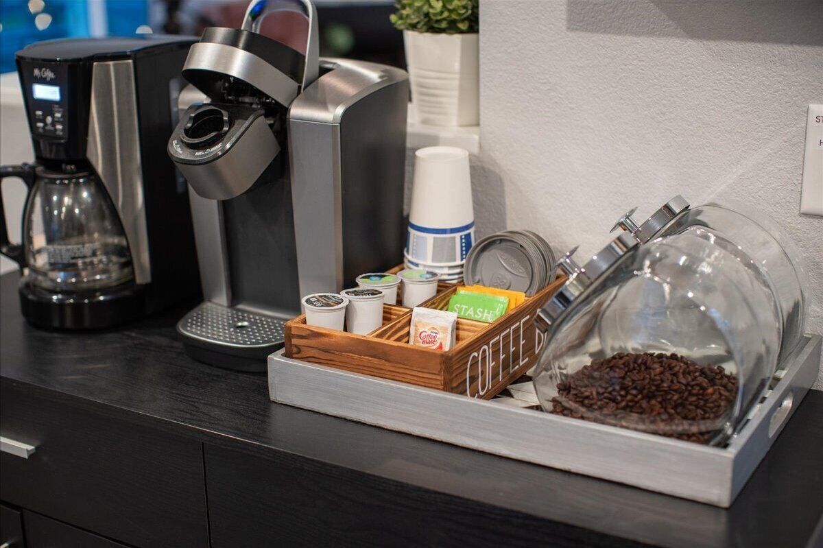 A coffee station with a coffee maker, a coffee dispenser, a glass container with coffee beans, and a wooden tray with coffee pods and a cup.