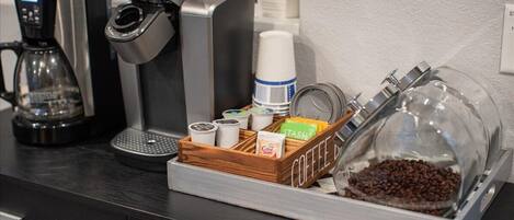 A coffee station with a coffee maker, a coffee dispenser, a glass container with coffee beans, and a wooden tray with coffee pods and a cup.