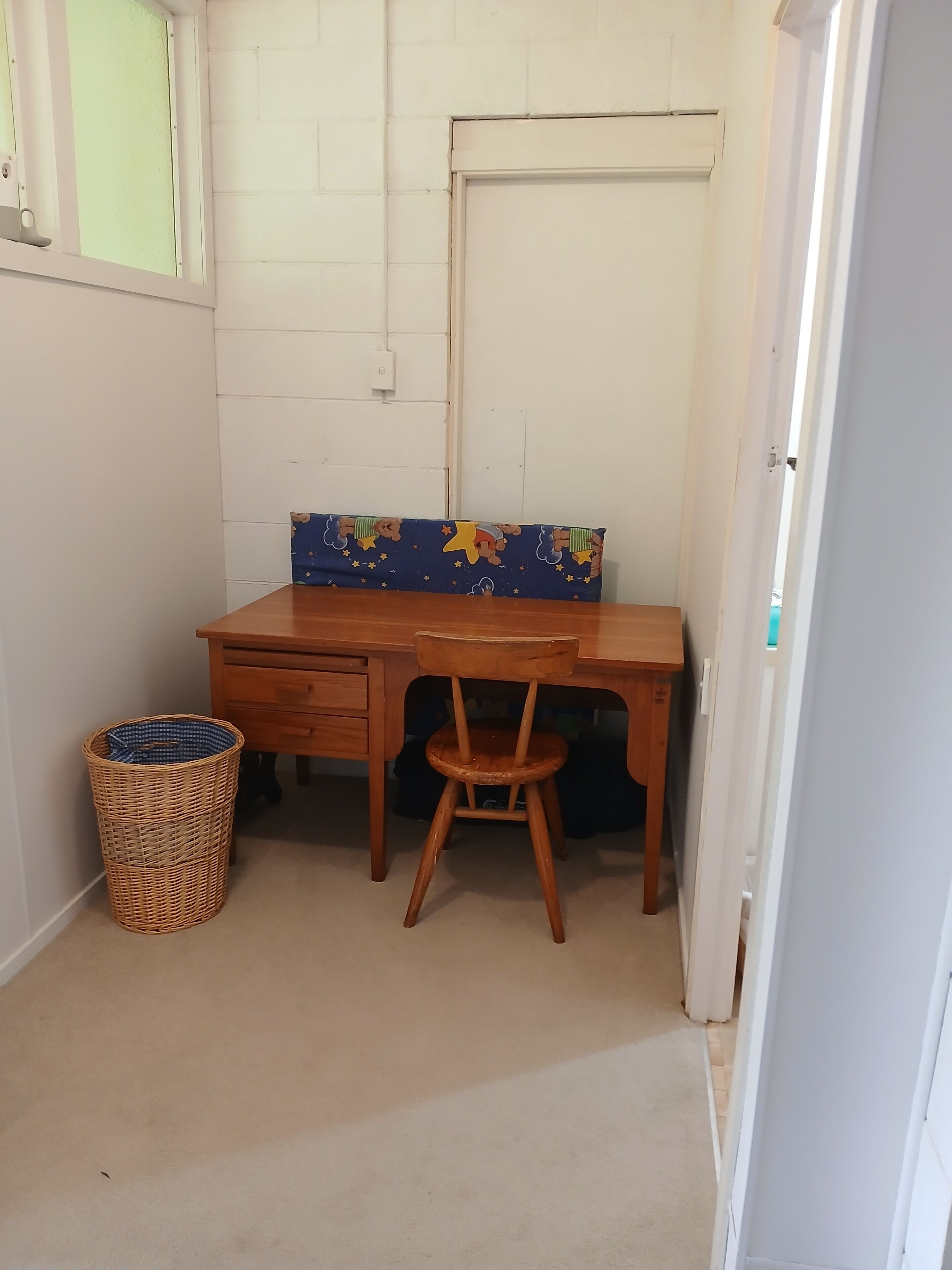Une petite pièce, bien rangée, avec un bureau en bois, une chaise, un panier en osier et un coussin bleu orné d’un motif d’ours.